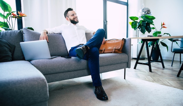 Confident stylish handsome bearded business man in glasses and formal smart wear is sitting on the couch in the modern office or room