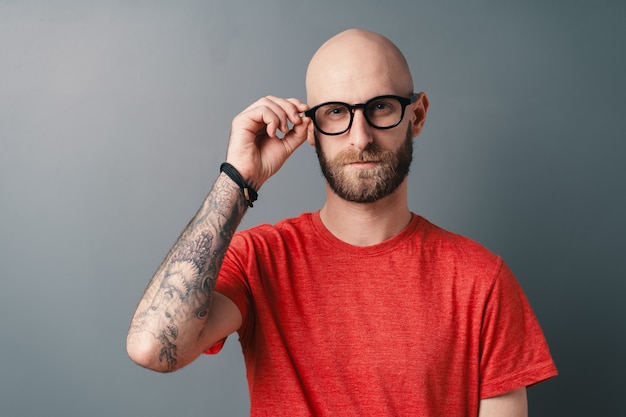 Foto fiducioso elegante giovane barbuto che fissa i suoi occhiali con cornice nera con la mano tatuata, in posa in studio su grigio.