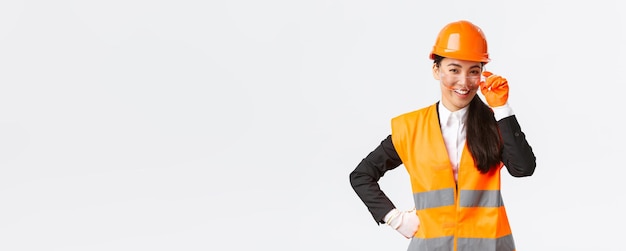 Уверенная в себе успешная азиатская женщина-главный менеджер гордо и улыбается в камеру в защитном шлеме, очках и перчатках на строительной площадке или на белом фоне предприятия