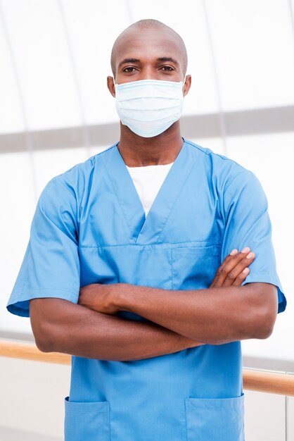 Confident and successful. Confident young African doctor in blue uniform and surgical mask looking at camera and keeping arms crossed