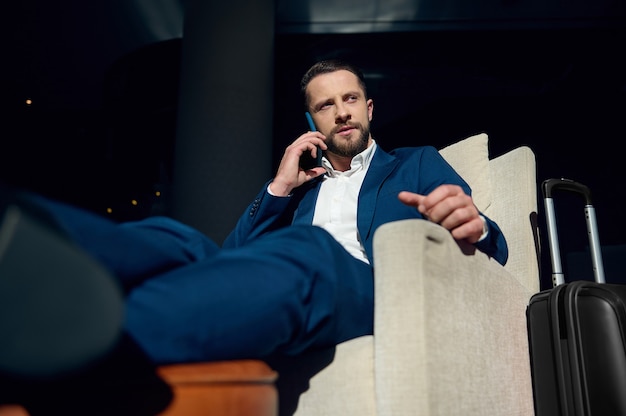Photo confident successful and perplexed young caucasian businessman talking on a mobile phone while sitting in an armchair of a hotel lounge, relaxing after a business trip. suitcase near armchair
