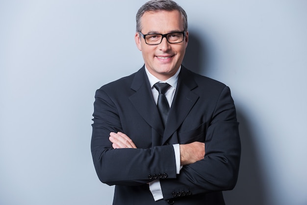 Confident and successful. Portrait of confident mature man in formalwear and eyeglasses looking at camera and smiling while standing against grey background