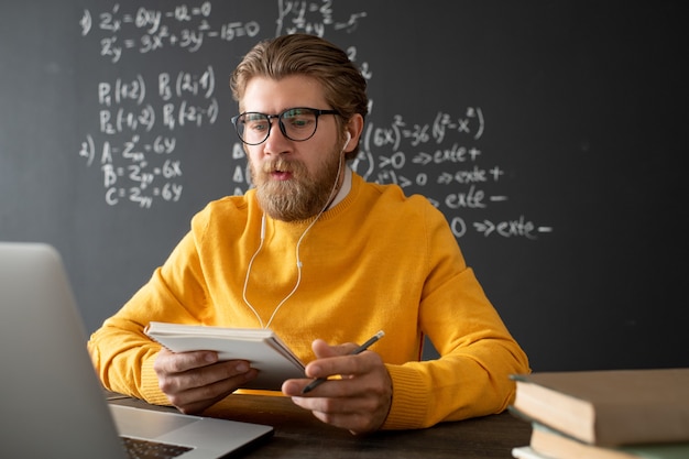 Confident teacher of algebra explaining new subject to his online students while sitting by table with books and notepad