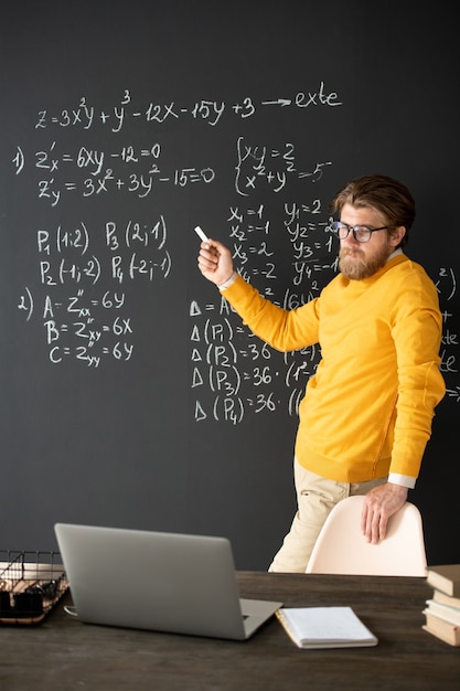 写真 オンラインレッスン中に生徒に説明しながら黒板の数式の1つを指しているチョークで自信を持って教師