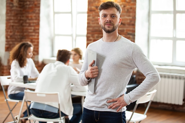 Confident team leader. Confident young man keeping arms crossed and looking at camera with smile while his colleagues working in the background.