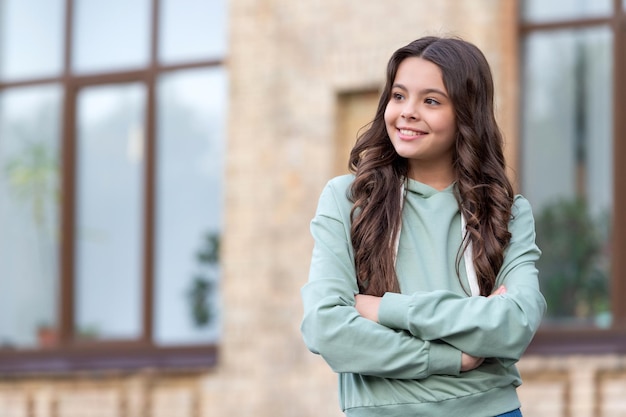 Confident teenage girl in casual hoodie looking aside keeping arms crossed blurry outdoors