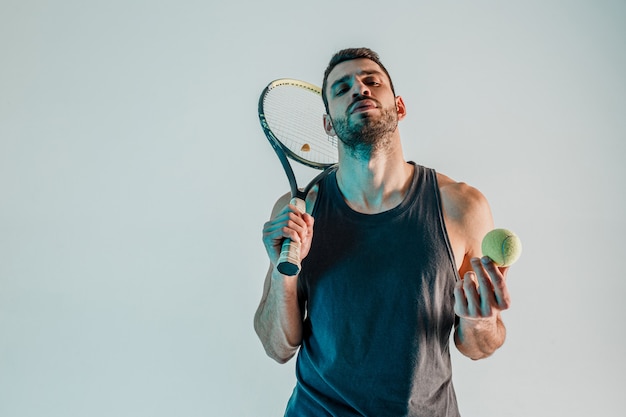 自信のあるテニスプレーヤーがボールとラケットを持っています。若いひげを生やしたヨーロッパのスポーツマンの正面図。ターコイズブルーの光で灰色の背景に分離。スタジオ撮影。コピースペース