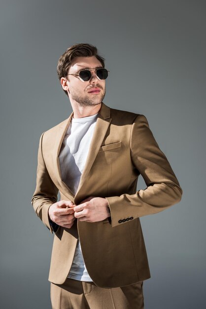 Confident trendy man in beige suite and sunglasses looking away on grey