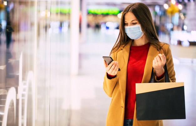 Fiduciosa donna alla moda in una mascherina medica di sicurezza con una borsa della spesa e smart phone sta camminando nel centro commerciale