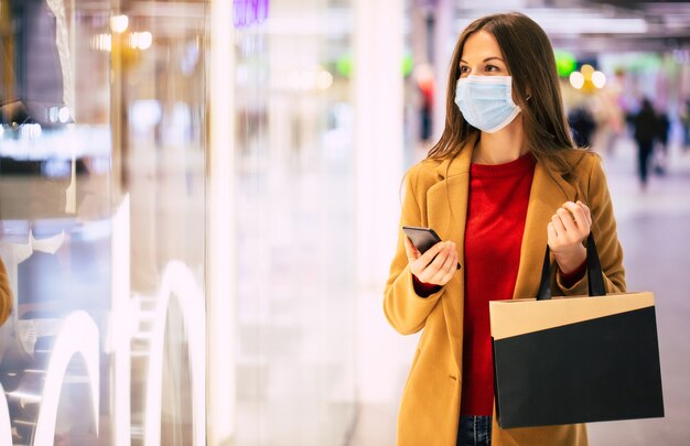 Уверенная в себе модная женщина в защитной медицинской маске с сумкой для покупок и смартфоном гуляет по торговому центру