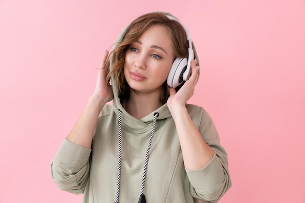 Уверенная женщина слушает музыку в наушниках Кавказская женщина наслаждается подкастом или аудиокнигами, одетая в негабаритную толстовку с капюшоном на розовом фоне крупным планом портрет