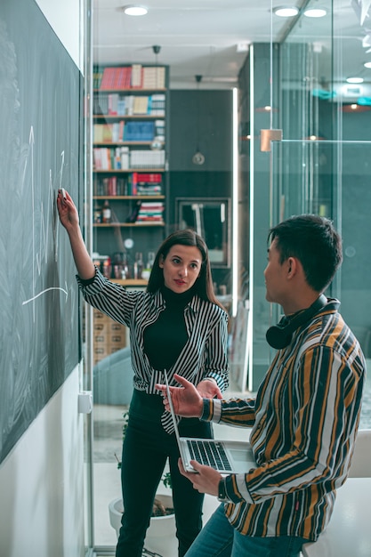 Confident woman standing with a piece of chalk near the blackboard and talking to her colleague