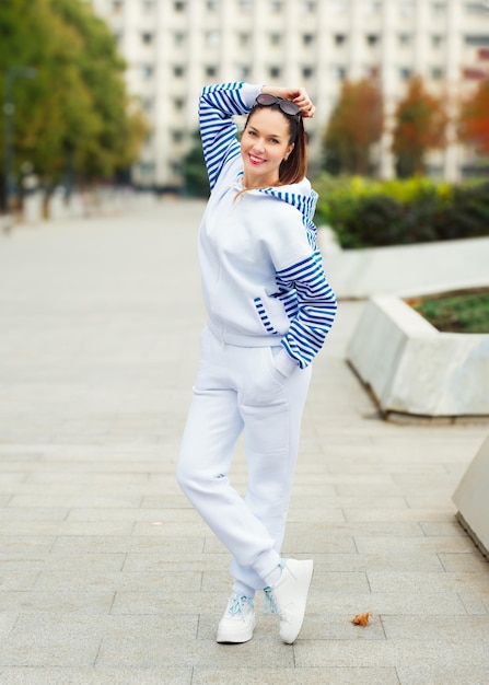 Photo confident woman in a stylish white tracksuit and bluestriped hoodie strikes a playful pose on a city promenade sunglasses on head