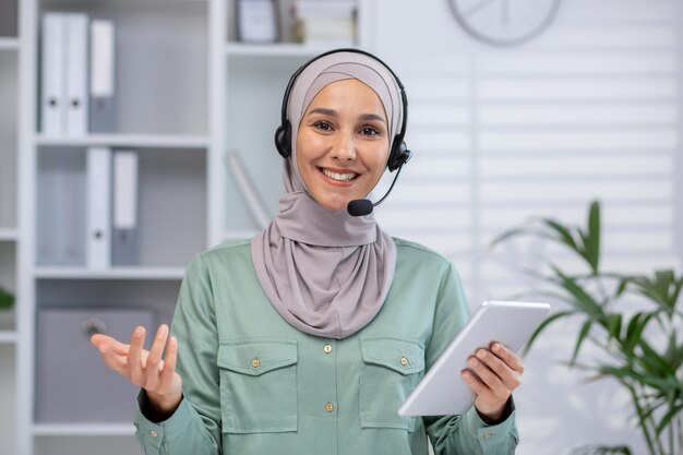 Уверенная в себе женщина в хиджабе и наушниках, работающая в современном офисе, она держит планшет и