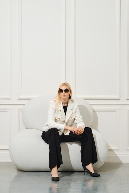 Confident woman in wide trousers blouse and woollen jacket sits in armchair with folded arms