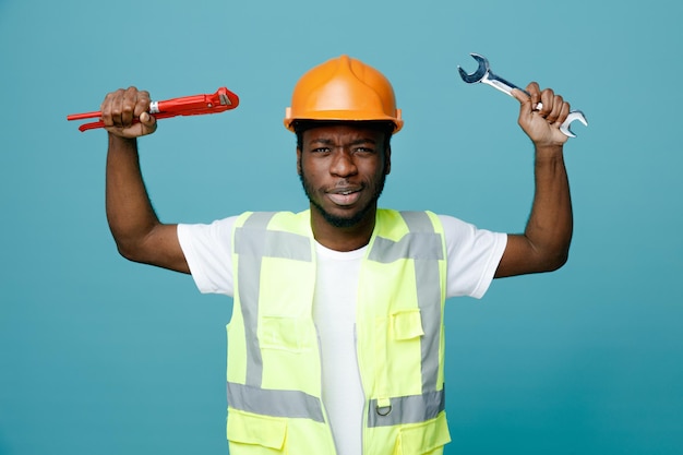 Confident young african american builder in uniform holding open end wrench with gas wrench isolated on blue background