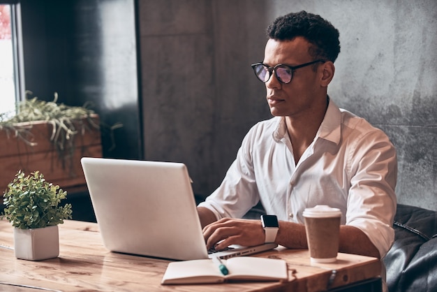 Photo confident young african man in smart casual wear using laptop while working in the office