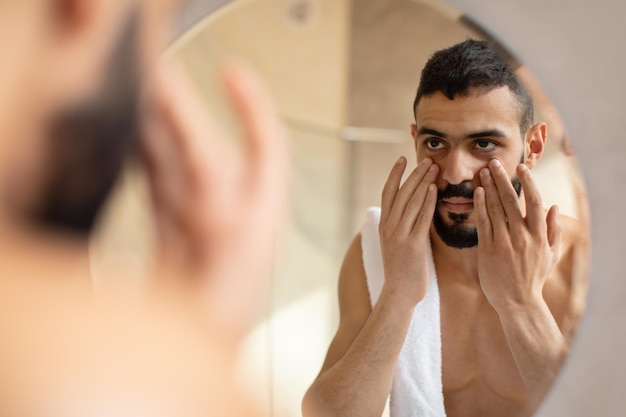 Confident young arab guy looking in the mirror applying cream