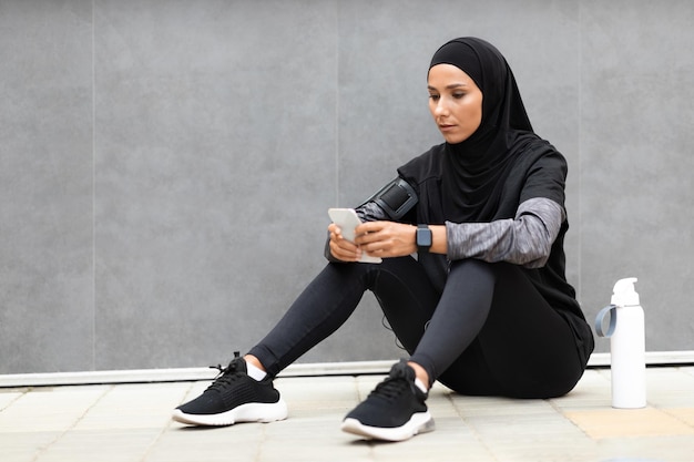 Confident young arab lady athlete in hijab sportswear with phone and water bottle sits on gray wall