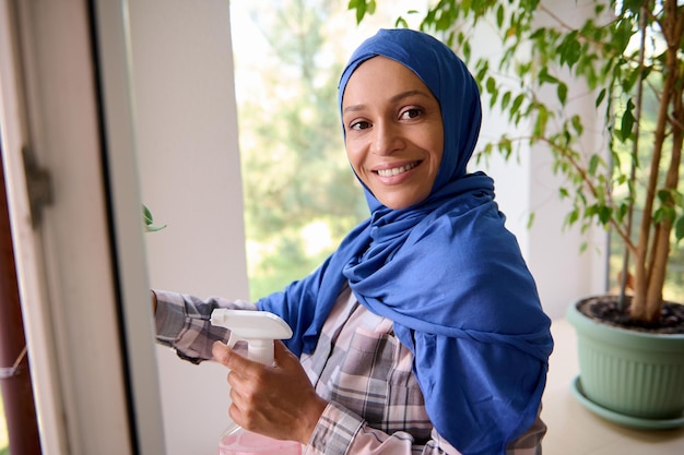 Confident young Arab Muslim pretty woman with covered head in blue hijab, smiles looking at camera, washing windows, removing stains and wiping with rag during spring cleaning in house. Copy ad space