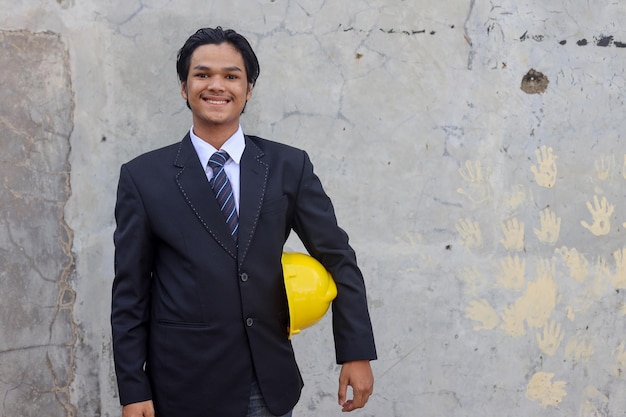 Confident young Asian engineer or businessman wearing black suit holding yellow helmet