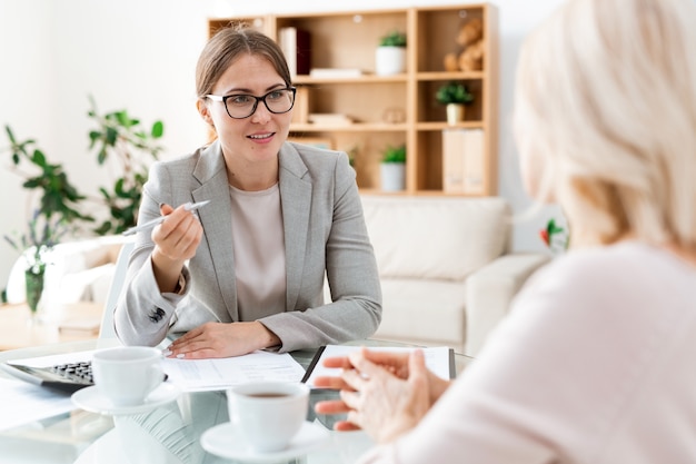 Confident young auditor with pen sitting in front of client and explaining one of points at meeting