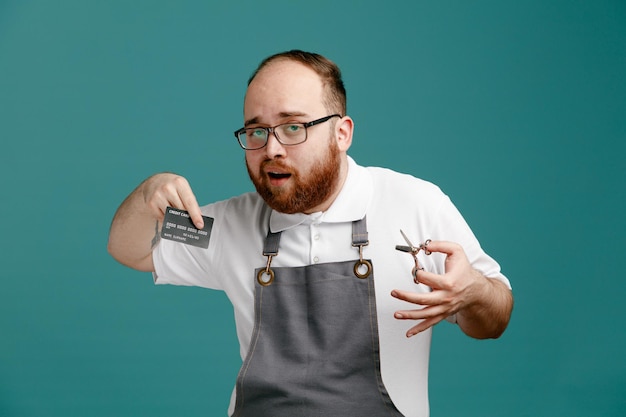 Confident young barber wearing uniform and glasses holding scissors looking at camera showing credit card isolated on blue background