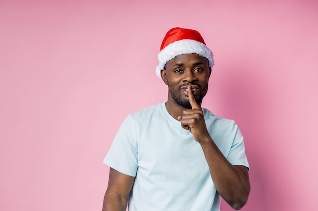 Confident young bearded afro american male with Santa hat on head, making shh gesture, keeping index finger on lips, telling secret, asking for silence, isolated over pink background.