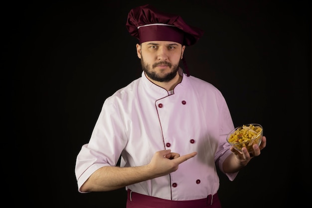 Уверенный молодой бородатый шеф-повар в униформе, указывая указательными пальцами на миску макарон в руке