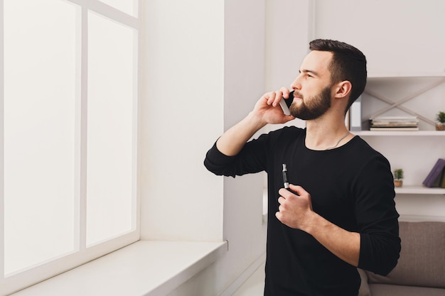 Confident young bearded man vaping electronic cigarette and talking on the mobile phone on white background. Vapor and alternative nicotine free smoking concept, copy space