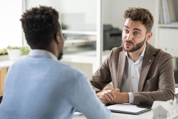 Confident young bearded recruiter sitting at table and asking candidate while holding job interview