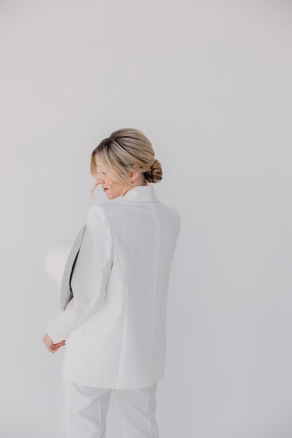 Confident young blond woman smiling looking at camera isolated on white background Studio portrait of successful friendly female in white suit and hat posing over white wall