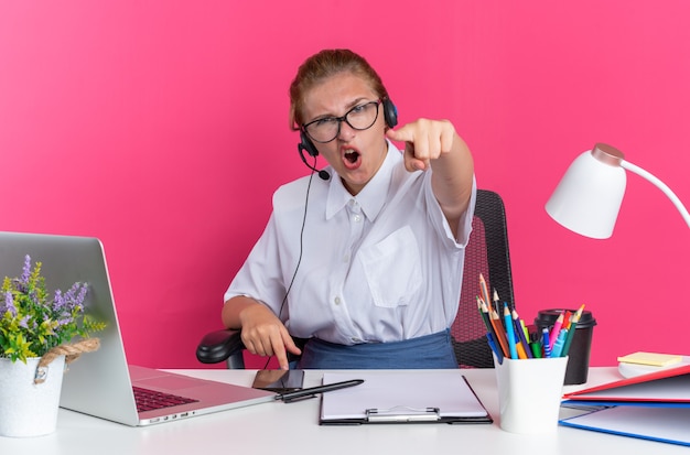 Confident young blonde call centre girl wearing headset and glasses sitting at desk with work tools looking and pointing at camera 