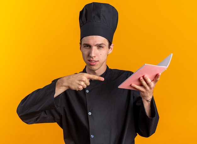 Confident young blonde male cook in chef uniform and cap holding and pointing at note pad looking at camera isolated on orange wall