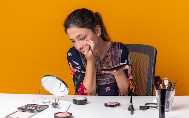 Confident young brunette girl sitting at table with makeup tools holding eyeshadow palette and applying eyeshadow with makeup brush looking at mirror 