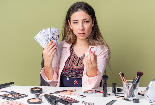 Confident young brunette girl sitting at table with makeup tools holding money and gesturing money sign isolated on olive green wall with copy space