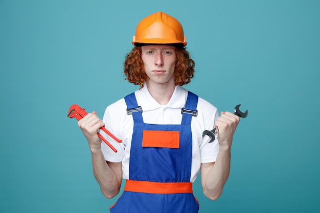 Confident young builder man in uniform holding wrenches isolated on blue background