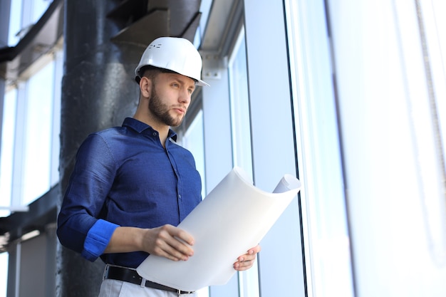 Confident young business man in shirt examining blueprint while standing against a window at office.