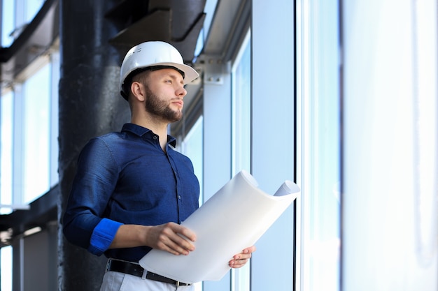 Confident young business man in shirt examining blueprint while standing against a window at office.