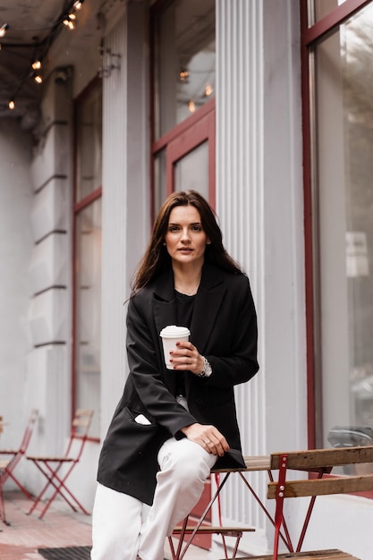 Confident young business woman with cup of coffee is posing outside of cafe on terrace Cheerful girl is weared casual business style is posing near cafe