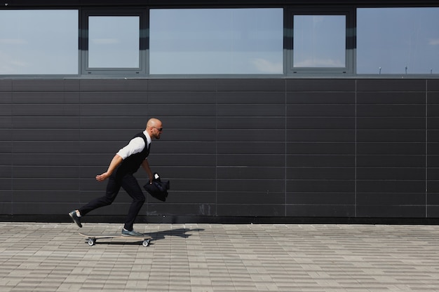 Confident young businessman in eyeglasses walking on the street, using longboard.