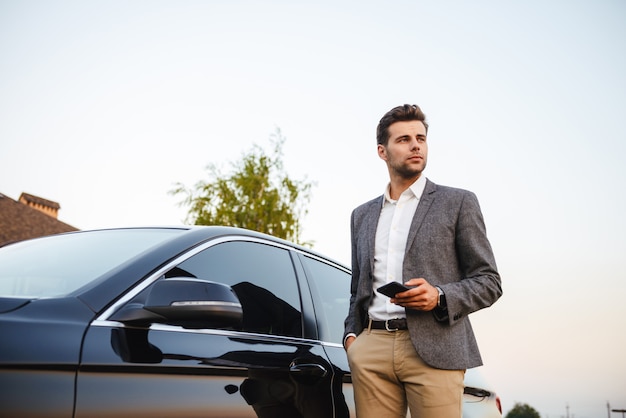 彼の車に立っているスーツで自信を持って若いビジネスマン