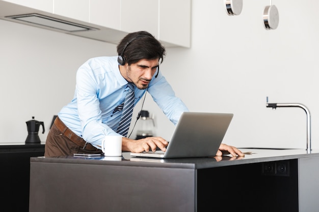 Confident young businessman working at home at the kitchen, using laptop computer