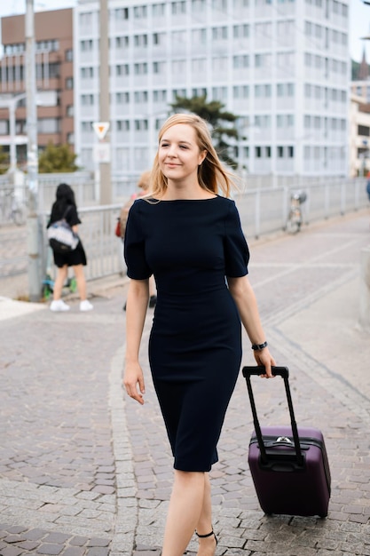 Confident young businesswoman with long blond hair in black dress carrying a suitcase in a city