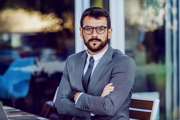 Confident young caucasian bearded businessman in suit and with eyeglasses sitting in cafe with arms crossed