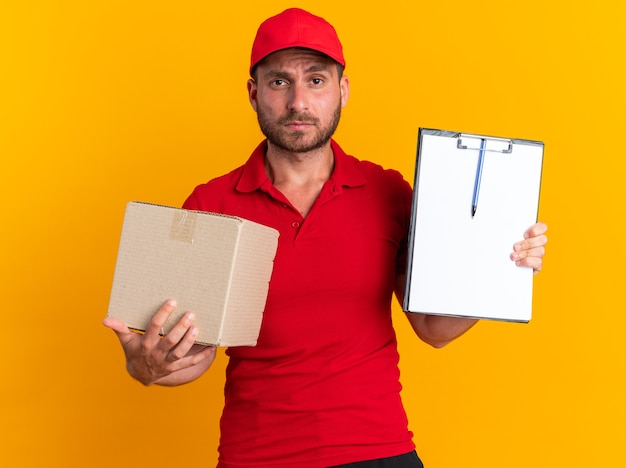 Confident young caucasian delivery man in red uniform and cap holding cardboard box looking at camera showing clipboard isolated on orange wall