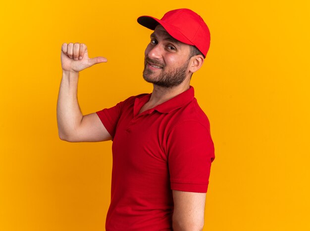 Confident young caucasian delivery man in red uniform and cap standing in profile view looking at camera pointing at himself isolated on orange wall with copy space