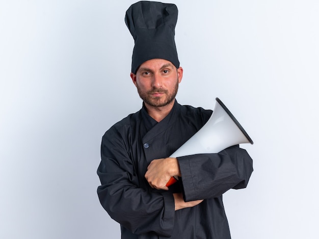 Confident young caucasian male cook in chef uniform and cap standing with closed posture looking at camera with speaker in hand isolated on white wall