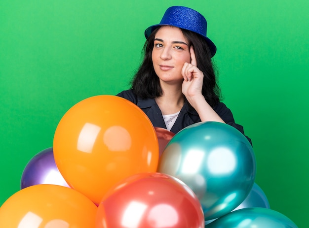 Confident young caucasian party woman wearing party hat standing behind balloons looking at front doing think gesture isolated on green wall