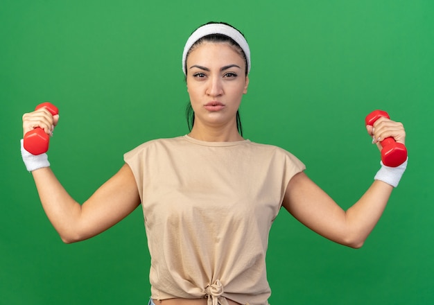 Confident young caucasian sporty girl wearing headband and wristbands raising dumbbells looking at front isolated on green wall